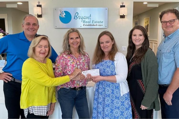 six people dressed in business casual pose smiling in front of Bryant Real Estate's sign in their office