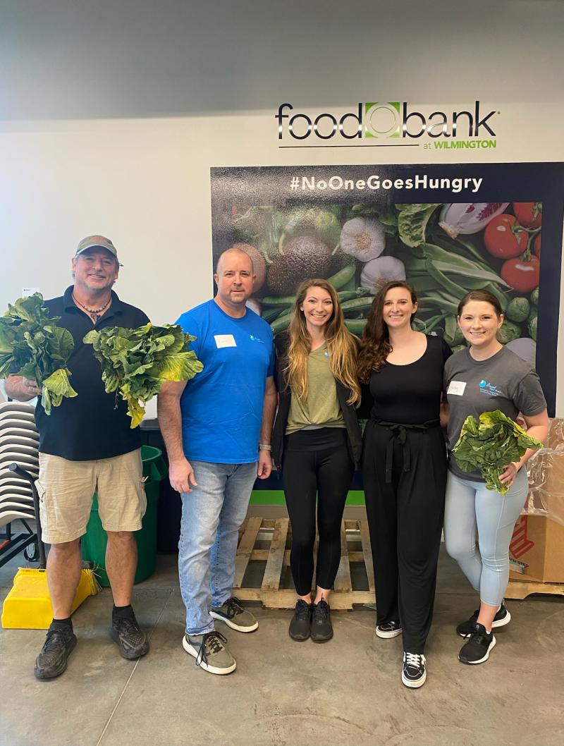 five people pose in front of a sign at a food bank. two of them hold heads of lettuce.